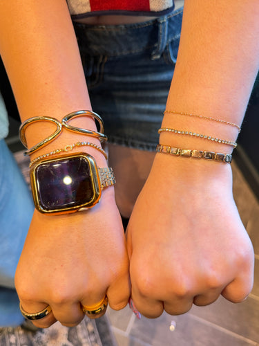 A close-up of a person's wrists wearing multiple gold-toned bracelets and rings, including a chunky geometric cuff, delicate chain bracelets, and a smartwatch with a gold case.