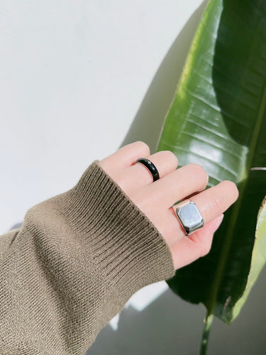 Hand wearing a brown sleeve with two rings, one black and one silver, against a green leaf background.